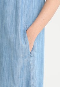 Vaaleansininen, chambray-kangasta oleva mekko, jossa on sivutasku, pehmeä rakenne ja rento muotoilu. Näkyvät ompeleet reunoilla.