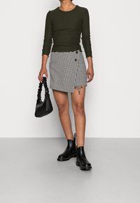 Olivenfarbenes geripptes Langarmoberteil, Rock im schwarz-weißen Hahnentrittmuster, schwarze Handtasche mit gedrehtem Griff und schwarze Ankle Boots.