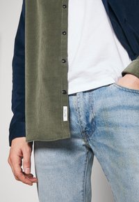 Vihreä corduroy-nappipaita, jonka hihat ovat tummansiniset, yhdistetty vaaleansinisiin farkkuihin; valkoinen t-paita alla, jossa näkyy paidan merkintä.