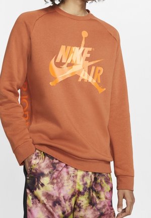 Person iført en brandsår-orange langærmet Nike Air sweatshirt og flerfarvede lilla, grønne og gule tie-dye bukser med den ene hånd i lommen.