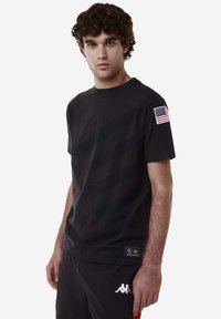 T-shirt nero a maniche corte con un motivo grafico sottile, caratterizzato da un patch della bandiera americana sulla spalla sinistra e un'etichetta di marca.