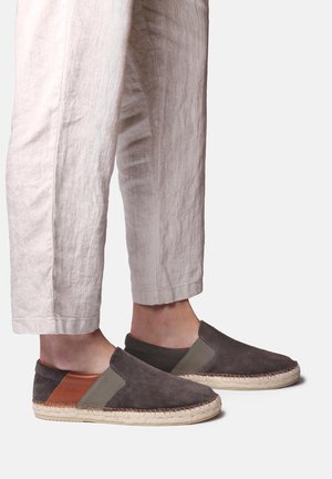 Nohy v tmavě šedých espadrilkách na nazouvání s béžovými provázkovými podešvemi a hnědými koženými záplatami na patě, doplněné světle béžovými zkrácenými kalhotami.