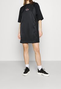 Osoba oblečená v černých šatech s krátkým rukávem a květinovým logem, s černo-bílými teniskami, stojící na lehkém jednobarevném pozadí.