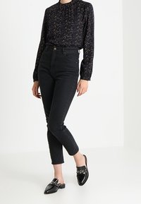 Jeans neri a vita alta abbinati a una camicia scura con motivo e mule nere con dettagli borchiati. Tessuto morbido, silhouette aderente.