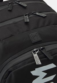 Sac à dos noir avec un tissu texturé, plusieurs compartiments zippés et un logo argenté bien en vue à l'avant. Le matériel comprend des zip décorés de la marque.