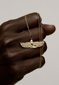 Collar de oro con un intrincado colgante alado que presenta grabados detallados, reposando sobre una mano oscura y texturizada con una cadena caída sobre los dedos.