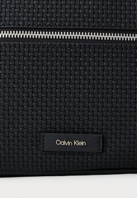 Μαύρη υφασμάτινη τσάντα με ασημένιο φερμουάρ, η οποία διαθέτει μια μικρή μαύρη δερμάτινη ετικέτα με ανάγλυφο το "Calvin Klein". Σατινέ επιφάνεια.