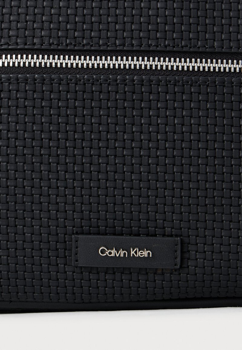 Borsa in tessuto intrecciato nera con cerniera argentata, dotata di un piccolo cartellino in pelle nera con "Calvin Klein" in rilievo. Superficie texturizzata.