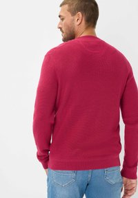 Maglione a girocollo lavorato a maglia di colore cremisi con maniche lunghe. Presenta un tessuto strutturato, polsini a coste e un design pulito sul retro. Indossato con jeans azzurri chiari.