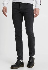 Jean slim noir en denim, avec un design à cinq poches, des passants de ceinture et des coutures visibles. Accentué par des chaussures noires à lacets.