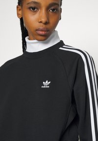 Čierna mikina s vysokým bielym golierom, ktorá má biele pruhy Adidas po pleciach a malé biele logo na hrudi.