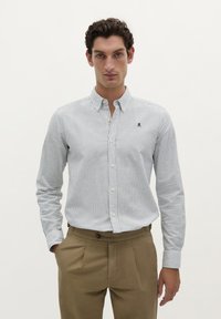 Camicia da uomo con bottoni, a righe orizzontali blu e bianche, in tessuto di cotone, con un logo sul petto, abbinata a pantaloni beige a pieghe.
