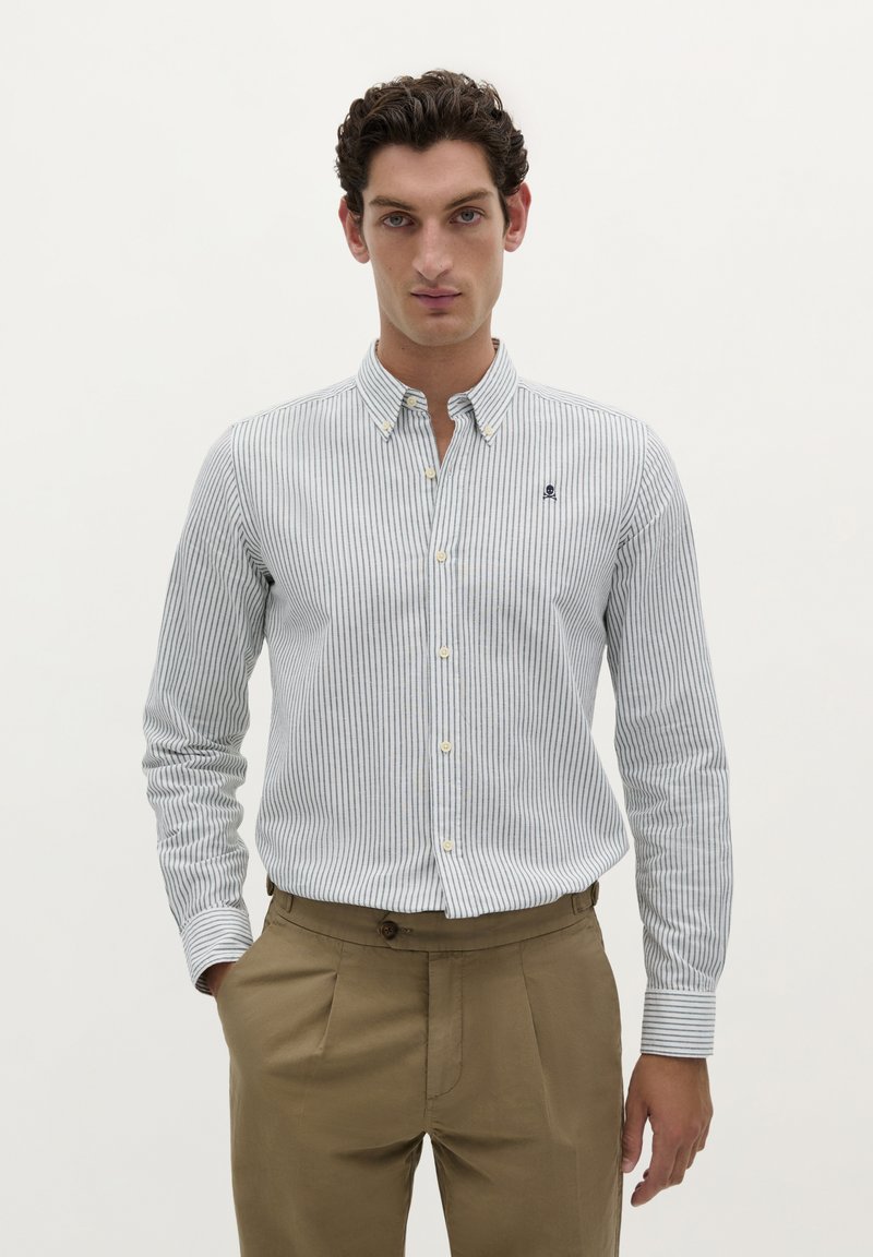 Camicia da uomo con bottoni, a righe orizzontali blu e bianche, in tessuto di cotone, con un logo sul petto, abbinata a pantaloni beige a pieghe.