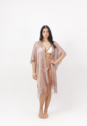 Vrouw die een witte bikini draagt en een doorschijnende, met franjes versierde lichtroze omslagdoek, blootsvoets staand tegen een witte achtergrond.