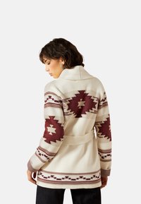 Strickjacke in Creme mit kastanienbraunen geometrischen Mustern, Gürtel in der Taille, Reverskragen und strukturierten Ärmeln.