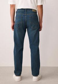 Sötétkék farmernadrág egyenes szabással, öt zsebbel és egy világosbarna folttal a hátsó derékrészen. Fehér sportcipővel viselve.