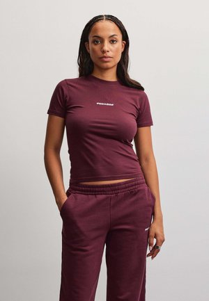 Donna con i capelli intrecciati che indossa una t-shirt bordeaux a maniche corte abbinata e pantaloni della tuta, in piedi con le mani nelle tasche davanti a uno sfondo semplice.