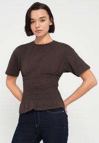 Καφέ βαμβακερό T-shirt με κοντά μανίκια, ζώνη με σούρες στη μέση και φαρδύ τελείωμα στο κάτω μέρος. Φοριέται με σκούρο μπλε τζιν, πάνω σε λευκό φόντο.