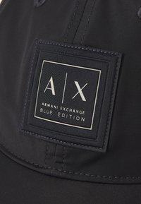 Fekete sapka sima anyagból, elöl egy négyzet alakú címkével, amelyen a fehér szöveg olvasható: "A|X ARMANI EXCHANGE BLUE EDITION".