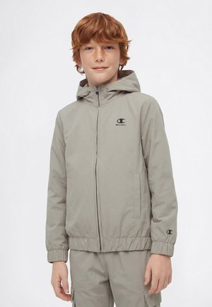 Rothaariger Junge trägt eine hellgraue Champion-Kapuzenjacke mit Reißverschluss und passende Cargohosen vor weißem Hintergrund.