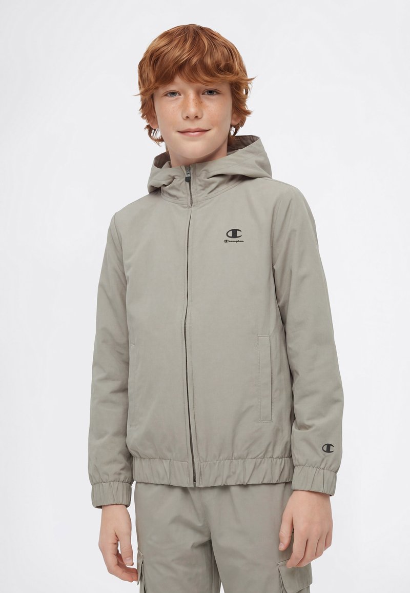 Rudy chłopiec w jasnoszarej bluzie Champion z kapturem na zamek błyskawiczny i dopasowanych spodniach typu cargo na białym tle.