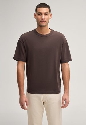 Mann steht mit Blick nach vorn, trägt ein schlichtes dunkelbraunes kurzärmeliges T-Shirt und hellbeige Hose vor neutralem Hintergrund.