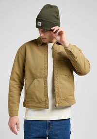 Bruine denim jas met een kraag en twee vozakken, gecombineerd met een groene gebreide beanie. Heeft een ritssluiting en zichtbare stikdetails.