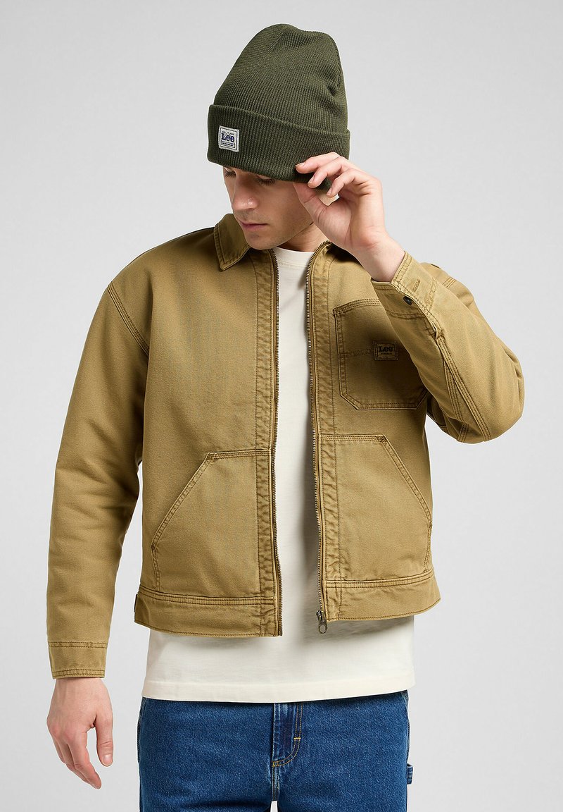 Bruine denim jas met een kraag en twee vozakken, gecombineerd met een groene gebreide beanie. Heeft een ritssluiting en zichtbare stikdetails.