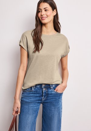 Frau mit langen braunen Haaren, die ein khakifarbenes kurzärmeliges Oberteil und blaue Jeans trägt, hält in einer Hand einen braunen Riemen, vor einem schlichten Hintergrund.