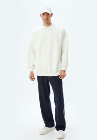 Sudadera blanca de gran tamaño con mangas largas, combinada con pantalones azul marino con franjas laterales y zapatillas blancas. Diseño sencillo y textura suave.