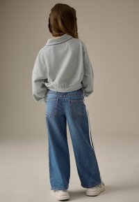 Sivý, skrátený, dlhý rukáv mikina v kombinácii s modrými širokými džínsami s bielymi bočnými pruhmi. Biely tenisky dotvárajú outfit.