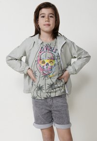 Giacca grigia chiaro con zip e cappuccio, sovrapposta a una maglietta grafica con un design colorato di teschio e palma, abbinata a pantaloni corti grigi arrotolati.