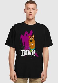 Schwarzes T-Shirt mit einem Cartoon-Hund und einem Geistermotiv, mit dem Text "BOO!" in fettgedruckten, weißen Buchstaben und Spinnennetz-Akzenten.