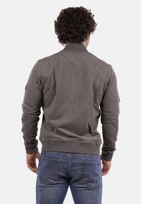 Giubbotto bomber grigio con colletto a costine, tessuto texture e dettagli delle cuciture. Presenta un piccolo logo sulla spalla sinistra. Indossato con jeans blu.