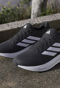 Paire de chaussures de course Adidas noires avec des bandes blanches et des semelles Cloudfoam placées sur un sol texturé en extérieur.