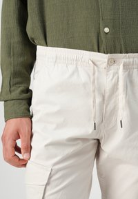Light beige elastic-waist pants with drawstring, side cargo pocket, and smooth texture, paired with a dark green textured shirt.