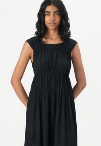 Svart ärmlös klänning med rynkad textur, rundad halsringning och empireliv. Tyget faller elegant och har sidofickor.