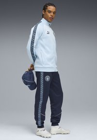 Helle blaue Zip-Jacke mit marineblauen Hosen, die gemusterte Seitestreifen und ein Logo aufweisen. Das Model hält eine marineblaue Mütze und trägt hellblaue Fußballschuhe.