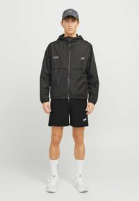 Junger Mann, der in einer schwarzen Kapuzenjacke, schwarzen Shorts, weißen Socken, weißen Turnschuhen und einer dunklen gemusterten Mütze vor einem einfarbigen Hintergrund steht.