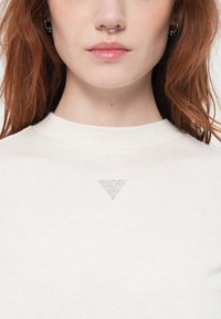 Žena s červenými vlasy, která má na sobě bílý svetr s kulatým výstřihem s malým trojúhelníkovým designem z kamínků umístěným uprostřed hrudníku.