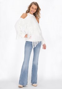 Witte gebreide trui met franjes, uitgesneden schouders, gecombineerd met lichtblauwe flared jeans en beige schoenen, geposeerd tegen een witte achtergrond.