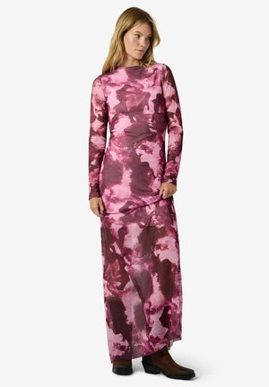 Une femme porte une robe à manches longues et jusqu’au sol, à motifs roses et bordeaux, sur un fond blanc uni.
