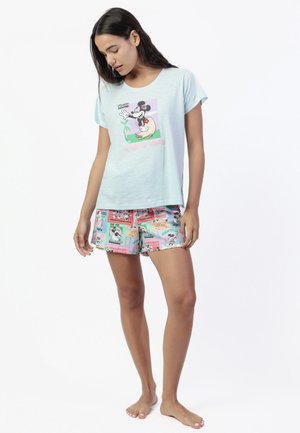 Mujer con camiseta azul clara de Minnie Mouse y pantalones cortos con estampado de dibujos animados coloridos, de pie descalza, mirando hacia abajo con postura relajada.