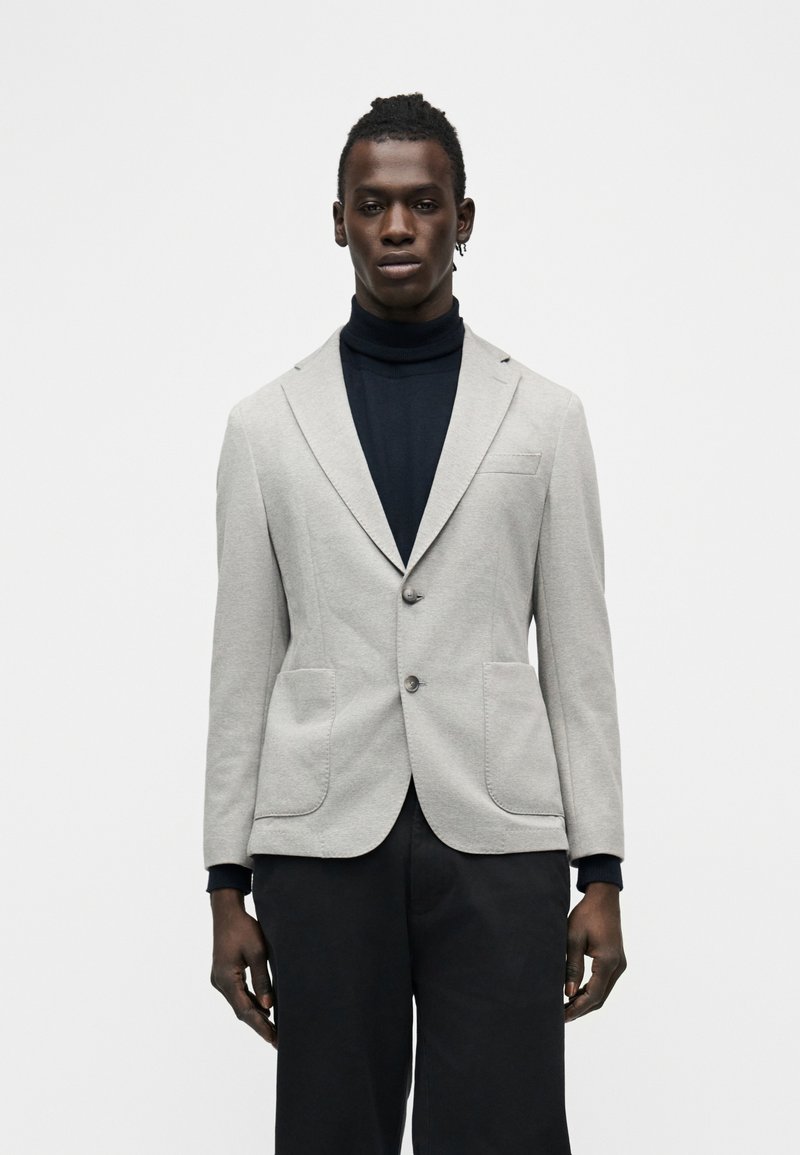 Άνδρας που φοράει ανοιχτό γκρι σακάκι πάνω από μαύρο πουλόβερ με ψηλό λαιμό και μαύρο παντελόνι, στέκεται μπροστά σε ένα απλό λευκό φόντο.