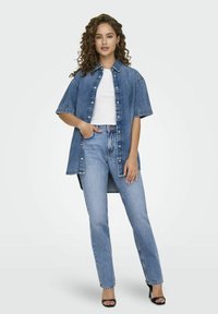 Νεαρή γυναίκα με σγουρά μαλλιά, φορώντας πολλαπλά επίπεδα τζιν πουκάμισο, λευκό μπλουζάκι, ανοιχτό μπλε τζιν και μαύρες γόβες με ανοιχτό δάχτυλο, στέκει με το ένα χέρι στην τσέπη.