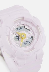 Reloj Casio Baby-G con una correa de goma rosa clara, pantalla digital con acentos amarillos y un fondo blanco que muestra el día y la hora.