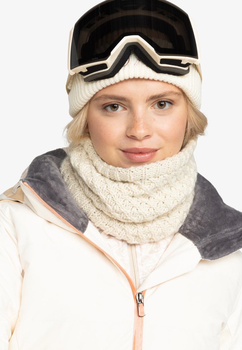 Bílý pletený šátek s texturovaným vzorem, spárovaný se světle barevnou lyžařskou bundou s šedým límcem a oranžovým zipem.