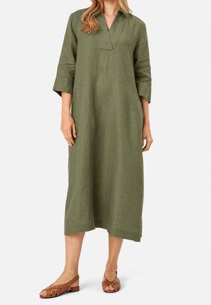 Vrouw die een olijfgroene midi-jurk met lange mouwen, V-hals en bruine gevlochten platte sandalen draagt.
