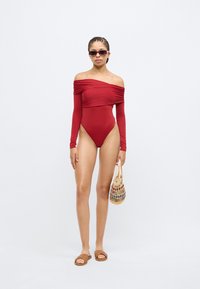 Punane õlgadeta ujumistrikoo, millel on pikad varrukad, kõrge lõikega jalgevahed ja kaeluse kokku kogutud detailid, kombineeritud pruunide sandaalide ja punutud kotiga.