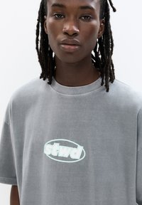 Graues, übergroßes T-Shirt aus weicher Baumwolle mit einem hellblauen "stwd"-Logo in ovaler Form auf der Vorderseite. Runder Ausschnitt, kurze Ärmel.
